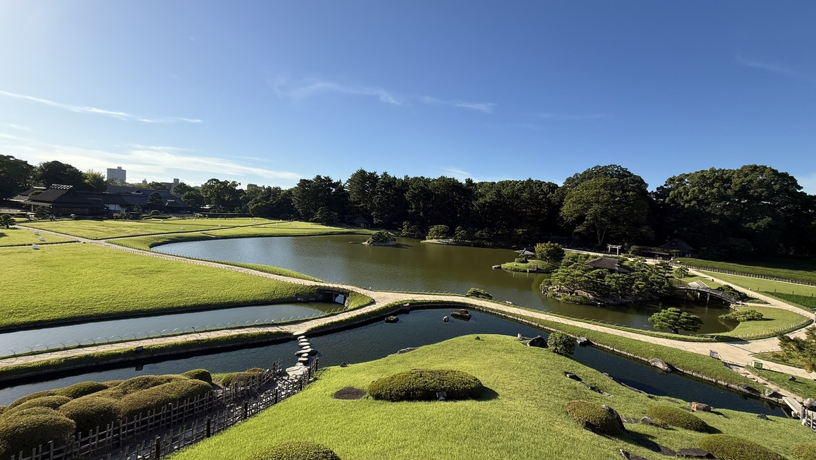 從後樂園水池遠眺，草丘與松樹後方，黑色的岡山城天守閣在藍天下清晰可見