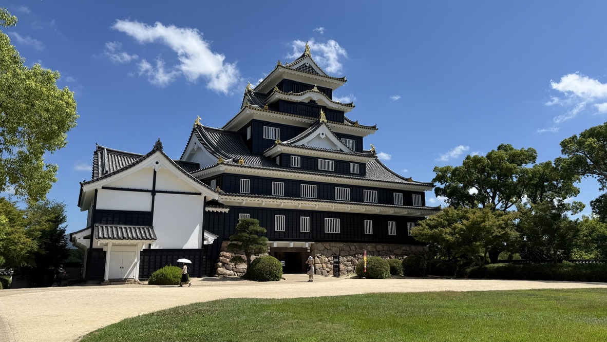 岡山城烏城全景,漆黑外牆在藍天白雲下格外醒目,入口前有遊客駐足仰望